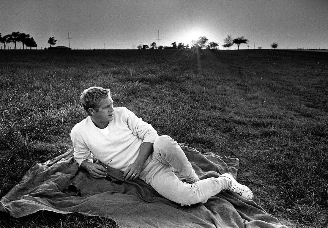Black and white photo of Steve McQueen lying on grass, wearing a white sweatshirt and white sneakers