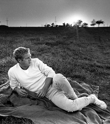 Black and white photo of Steve McQueen lying on grass, wearing a white sweatshirt and white sneakers