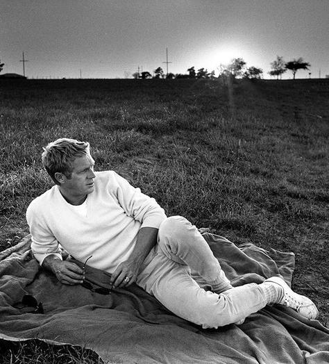 Black and white photo of Steve McQueen lying on grass, wearing a white sweatshirt and white sneakers