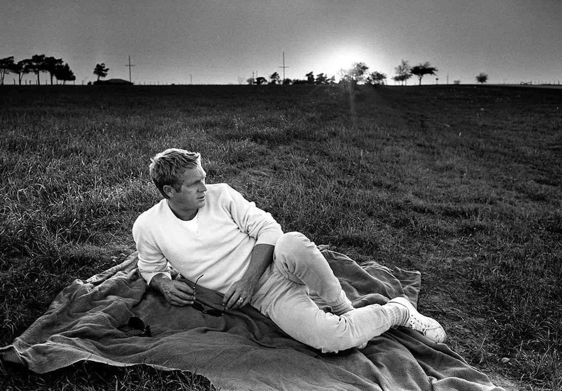 Black and white photo of Steve McQueen lying on grass, wearing a white sweatshirt and white sneakers
