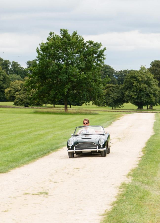 Small classic car driving up the driveway with a red helicopter coming in to land