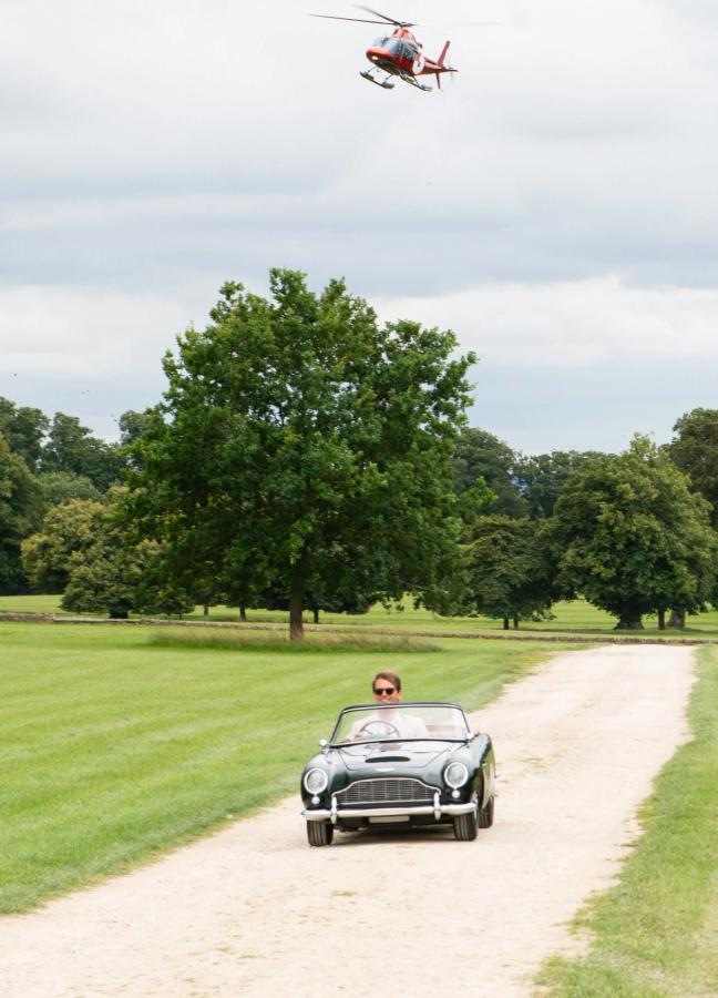 Small classic car driving up the driveway with a red helicopter coming in to land