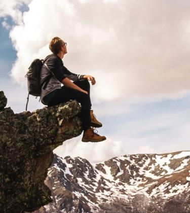 man sits on a crag on a mountain wearing boots and looking at the snow and sky