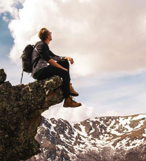 man sits on a crag on a mountain wearing boots and looking at the snow and sky