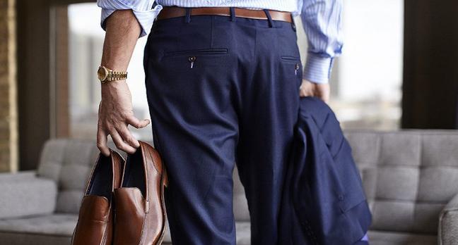 A man wearing navy suit trousers and brown belt, holding brown leather shoes