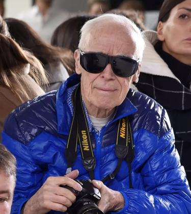 bill cunningham at fashion week, wearing sunglasses, iconic blue jacket and nikon DSLR camera