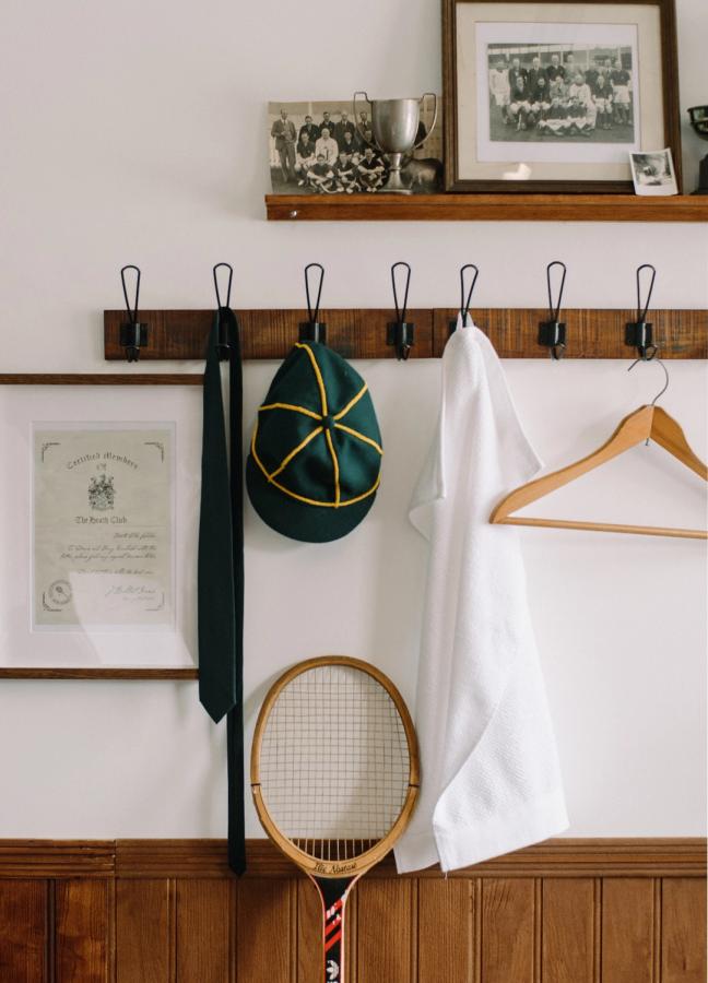 Changing room coat hooks with a tie, sun hat and white shirt tennis racket hung up. A shelf with old sports team photos, certificate, trophies and a tennis racket against the wall.