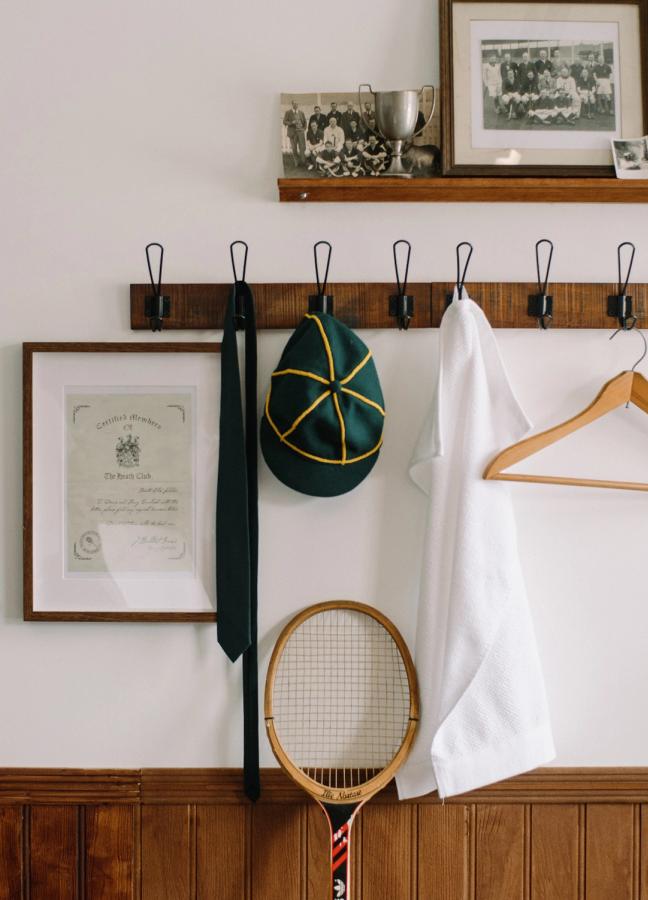 Changing room coat hooks with a tie, sun hat and white shirt tennis racket hung up. A shelf with old sports team photos, certificate, trophies and a tennis racket against the wall.