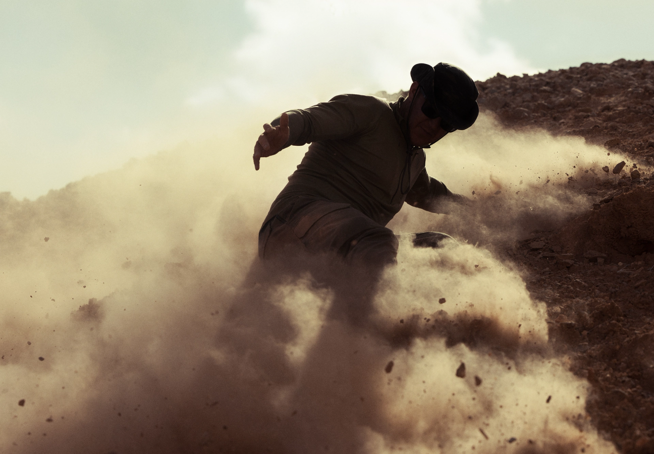 Man sliding down hill of loose rubble through dust clouds wearing a windguard jacket, sunglasses and watchman boonie hat