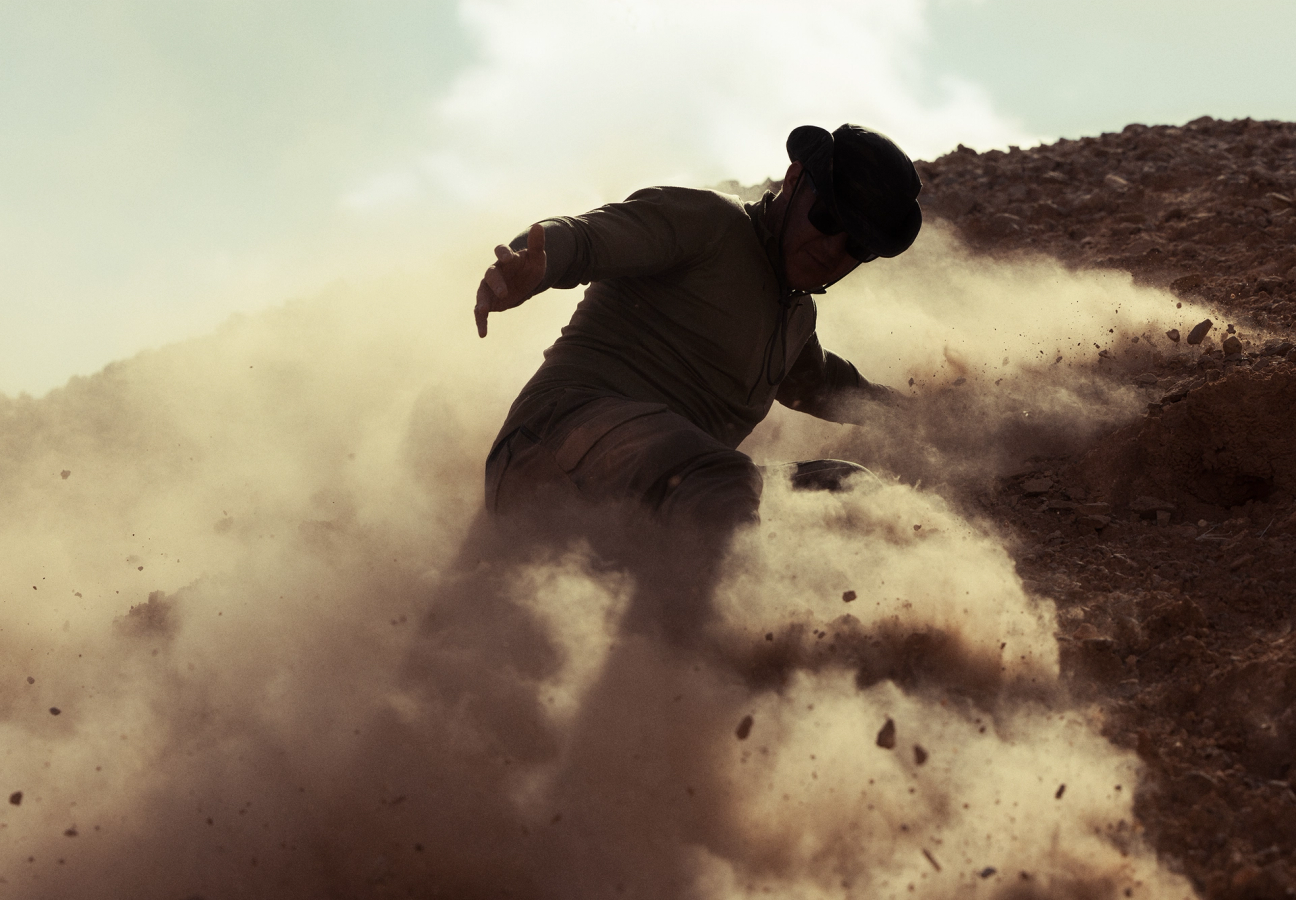 Man sliding down hill of loose rubble through dust clouds wearing a windguard jacket, sunglasses and watchman boonie hat