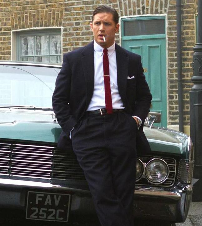 Tom Hardy sat on the bonnet of a car, wearing a suit balancing a cigarette in his mouth.