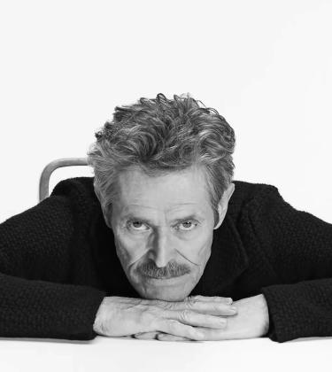 An image of actor Willem Dafoe wearing black and looking into the camera.