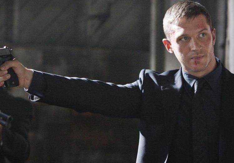 Tom hardy in an all black suit with gun in right hand held out at arms length