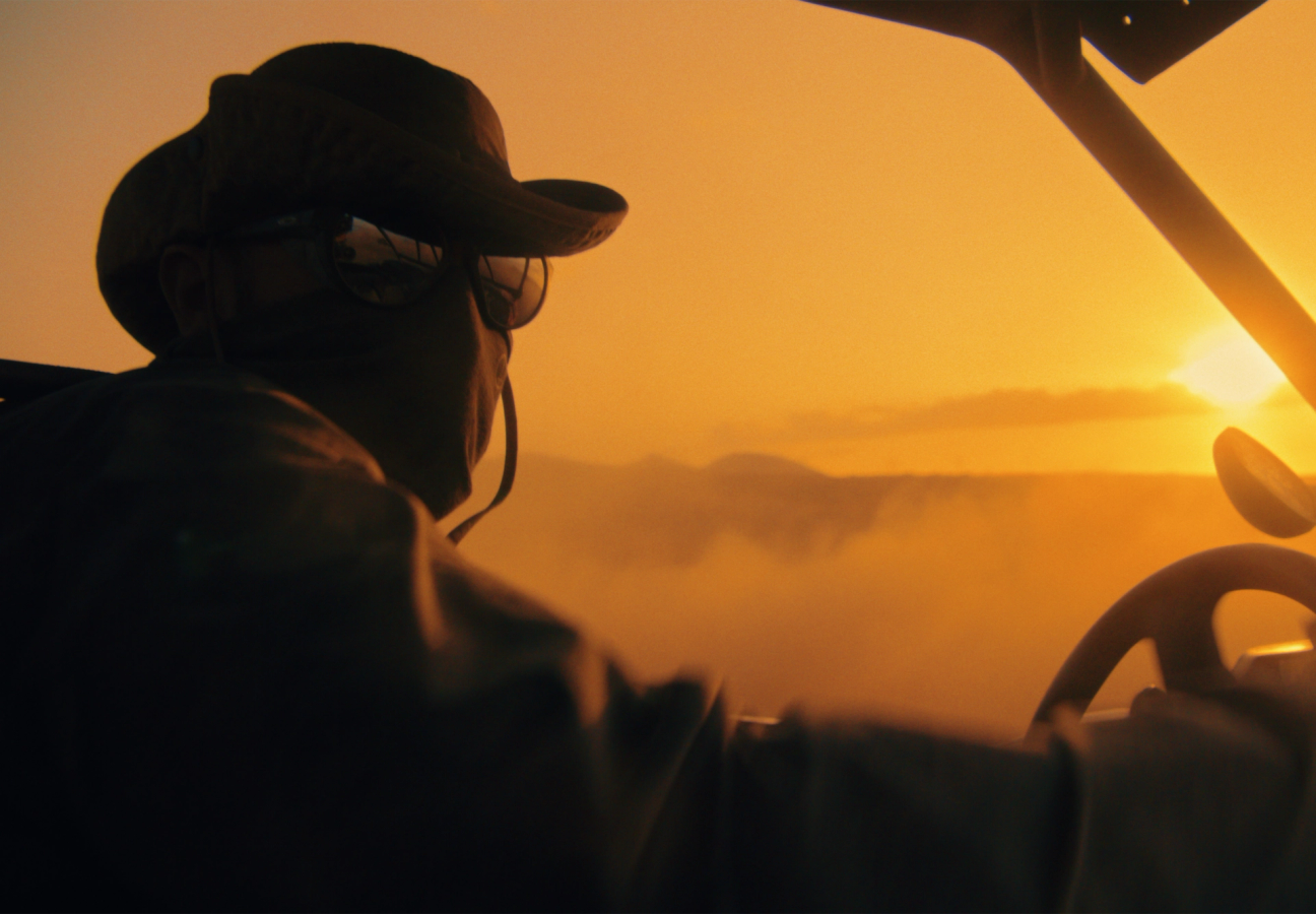 Man driving through a dusty landscape in a windowless vehicle wearing a windguard jacket, neck gaiter, sunglasses and watchman boonie hat