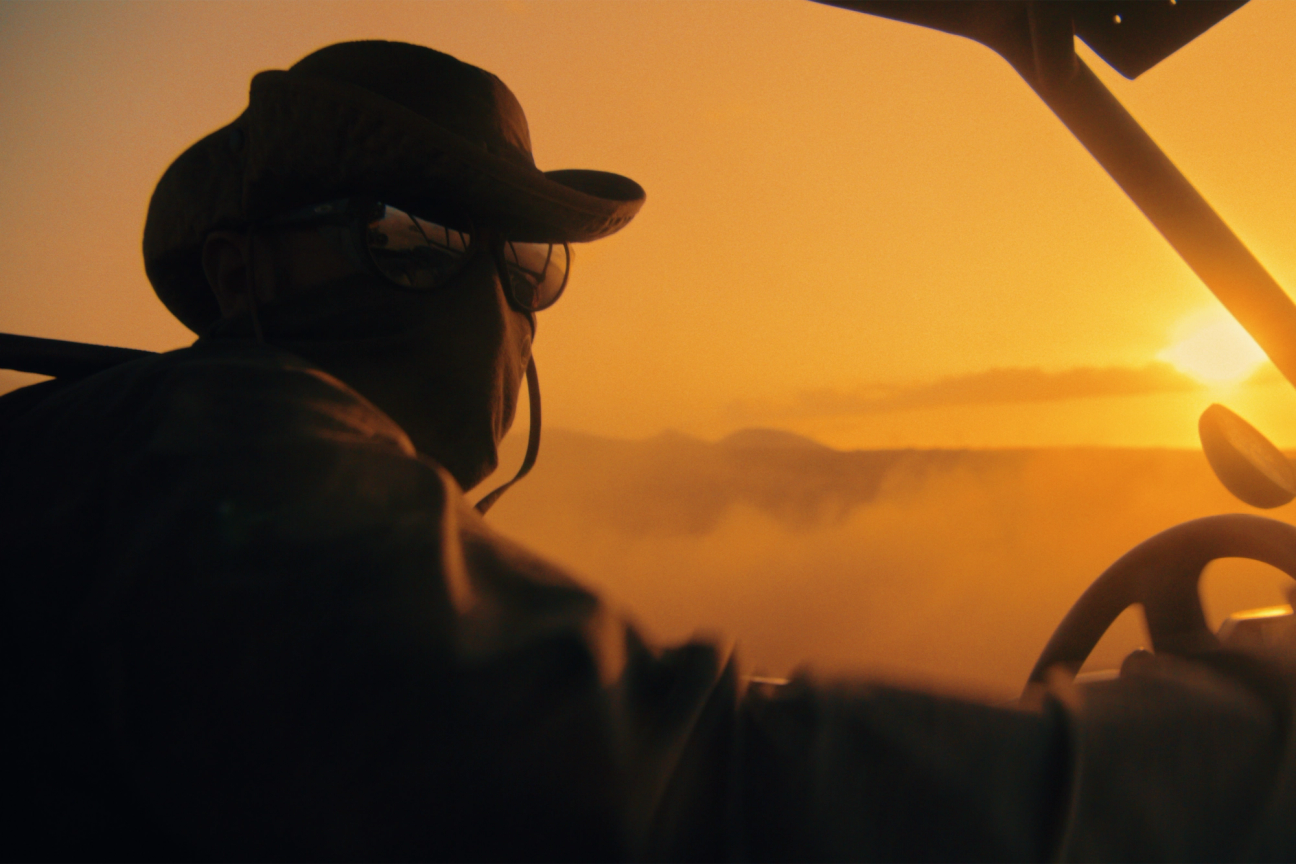 Man driving through a dusty landscape in a windowless vehicle wearing a windguard jacket, neck gaiter, sunglasses and watchman boonie hat