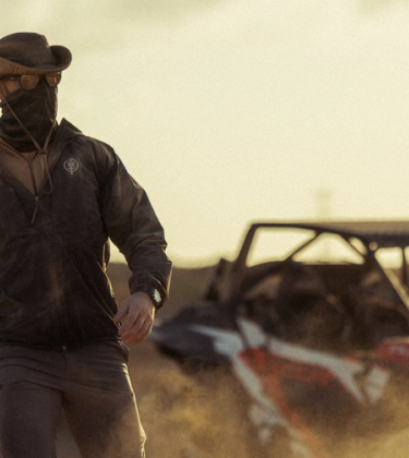 Man walking through a dusty landscape wearing a windguard jacket, oryon carbon trousers, neck gaiter, sunglasses and hat