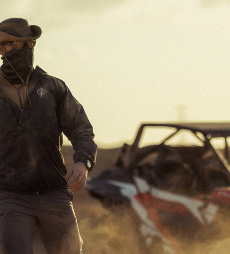 Man walking through a dusty landscape wearing a windguard jacket, oryon carbon trousers, neck gaiter, sunglasses and hat