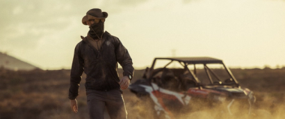Man walking through a dusty landscape wearing a windguard jacket, oryon carbon trousers, neck gaiter, sunglasses and hat