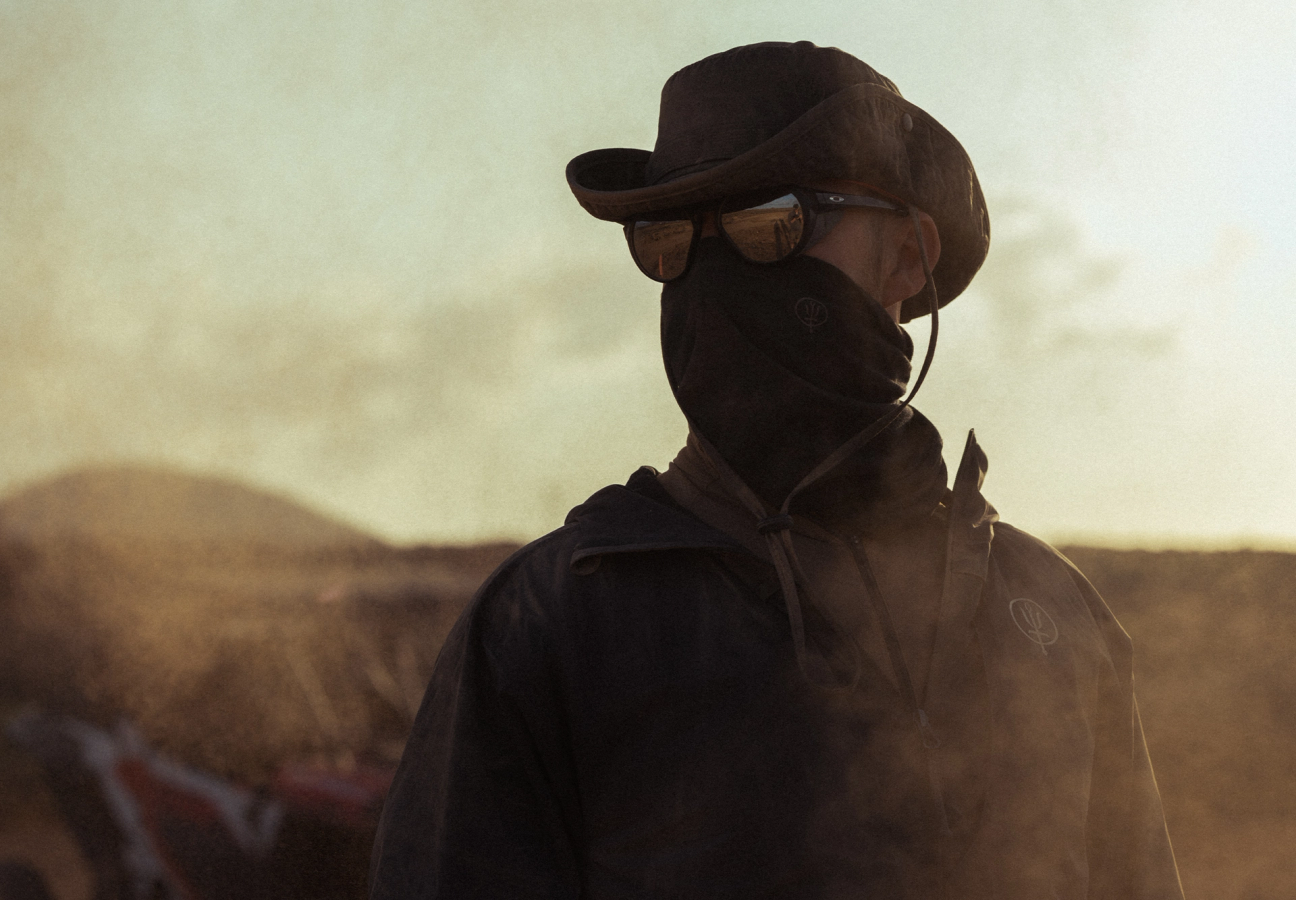 Man in a dusty landscape wearing a windguard jacket, neck gaiter, sunglasses and watchman boonie hat