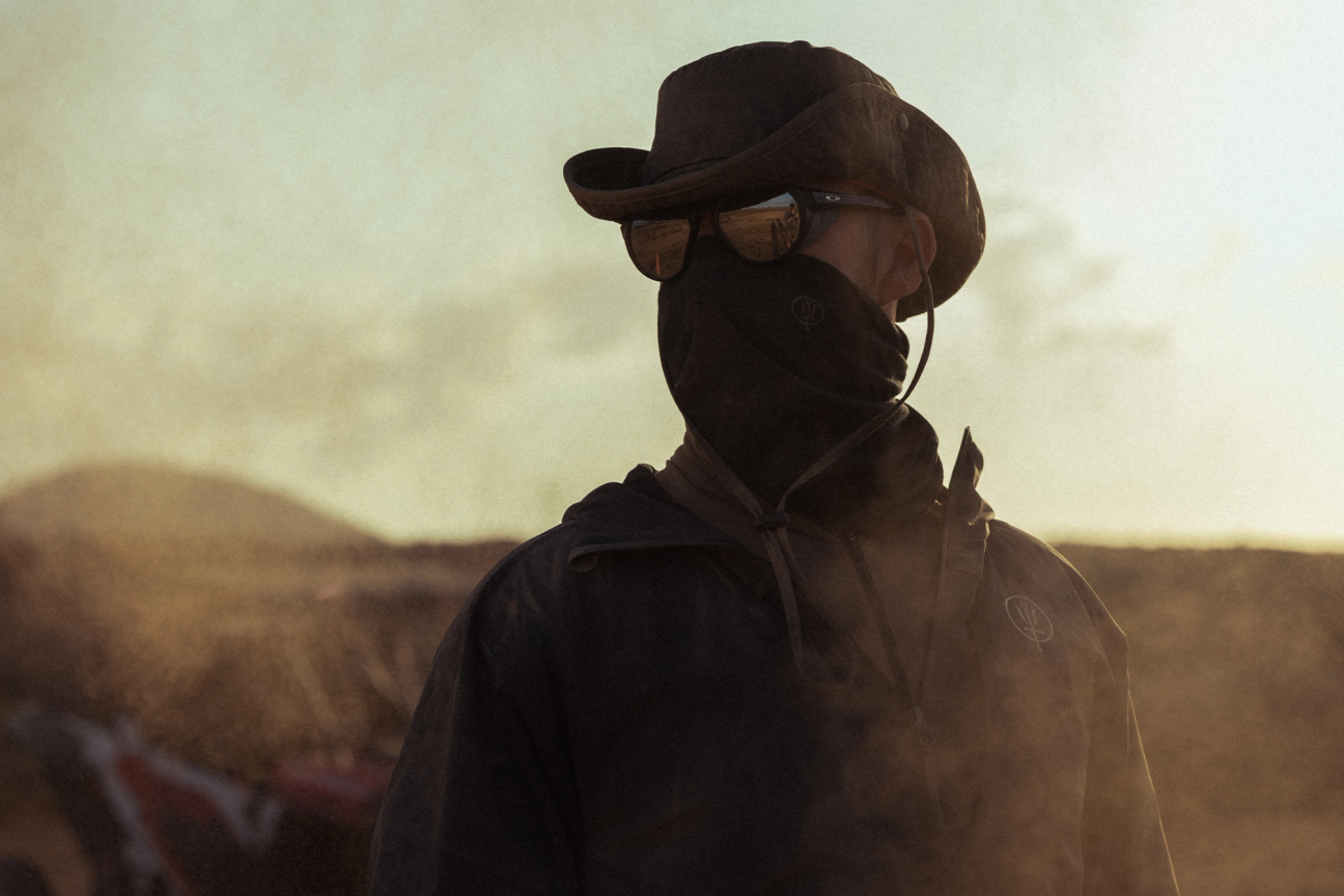 Man in a dusty landscape wearing a windguard jacket, neck gaiter, sunglasses and watchman boonie hat