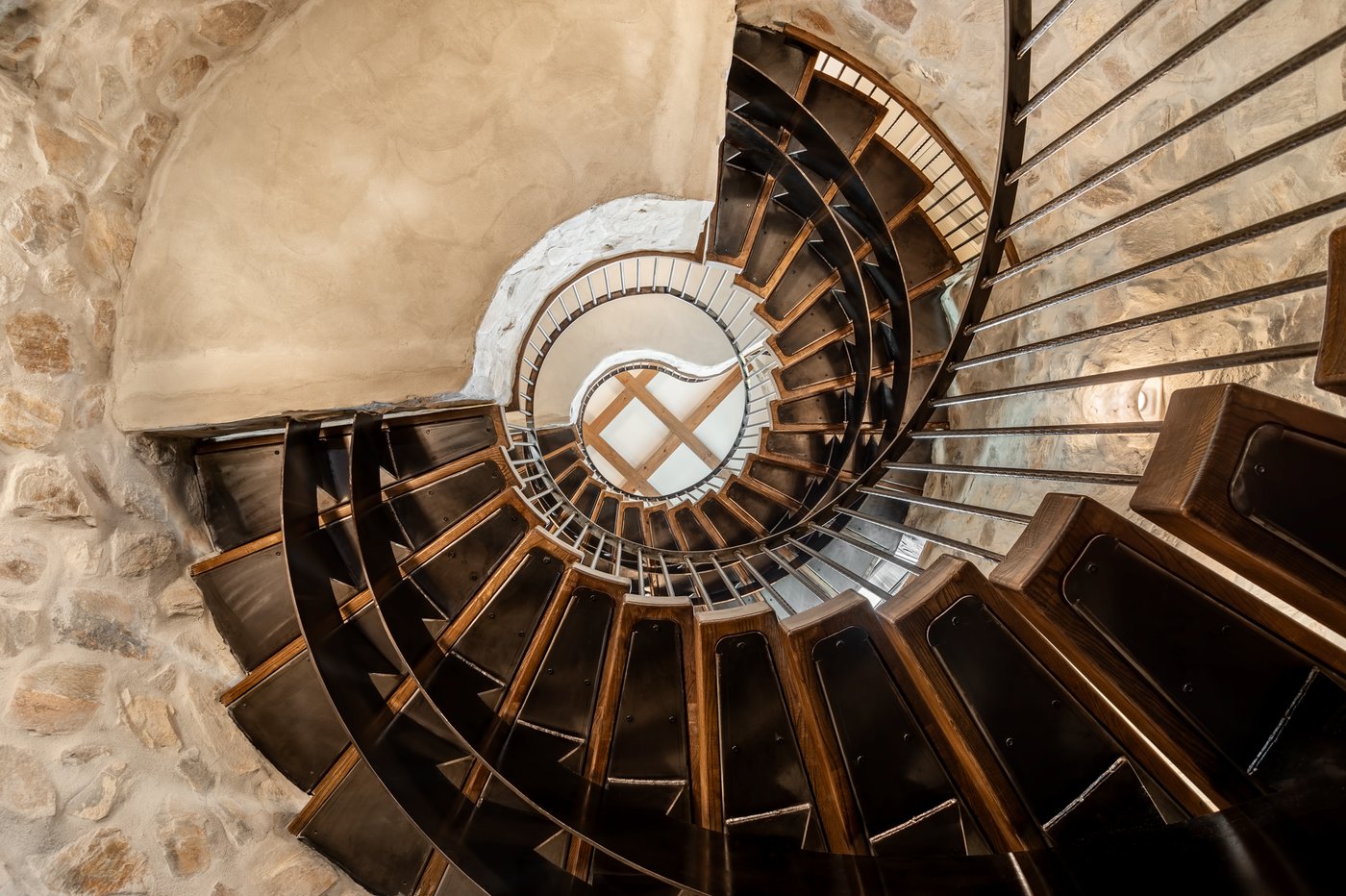 Looking down a spiral staircase with dark wood treads beside a stone wall.