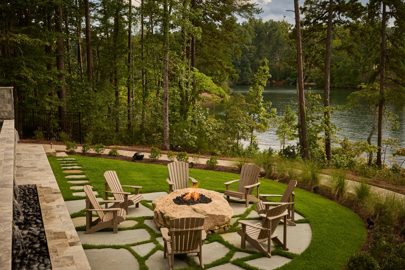 Adirondack chairs arranged around a round stone fire pit on a lakeside lawn.