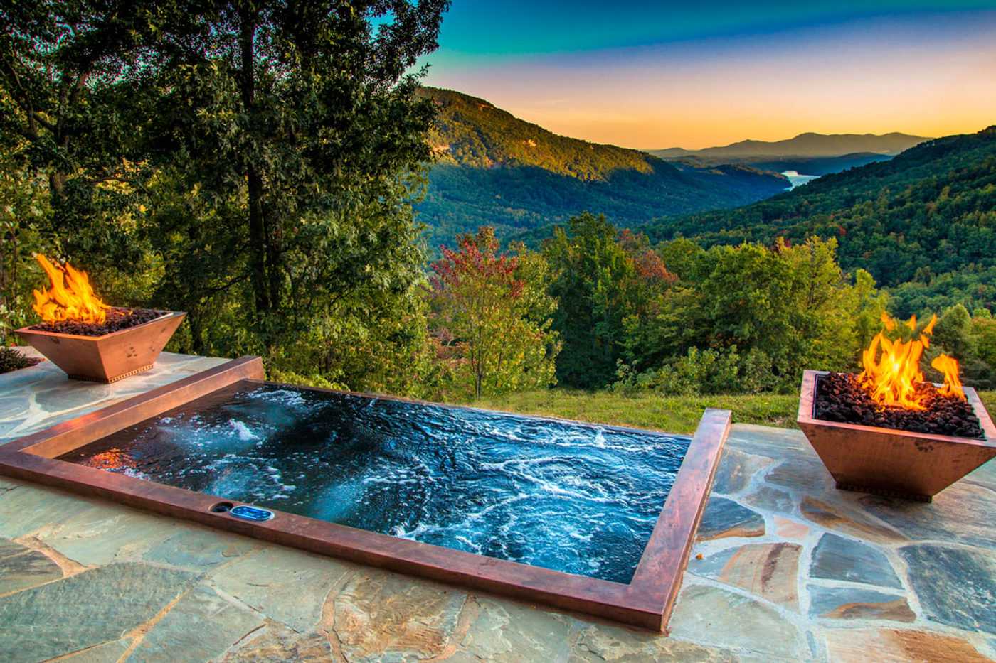 A hot tub on a stone patio flanked by two fire bowls, overlooking mountains at sunset.