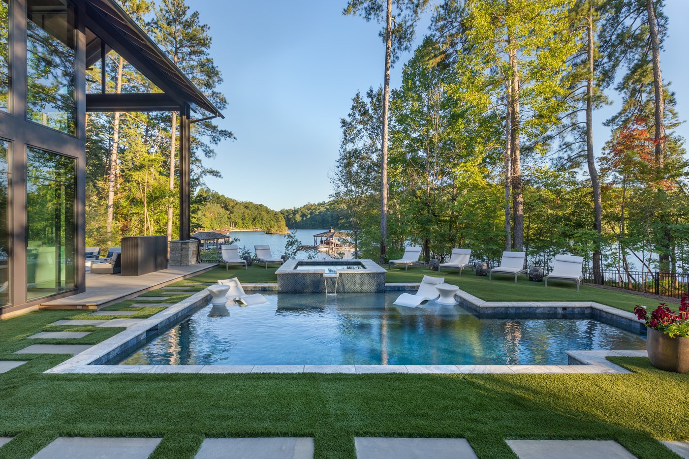 A pool and spa beside a modern lakeside home, with a lawn, loungers and the lake beyond.