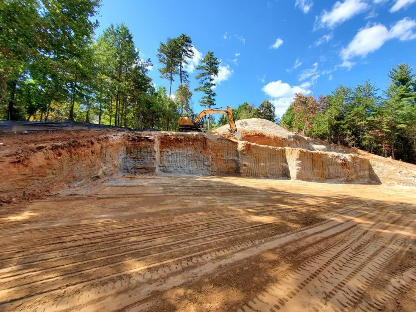 An excavator on a freshly graded red-clay building site cut into a wooded hillside.