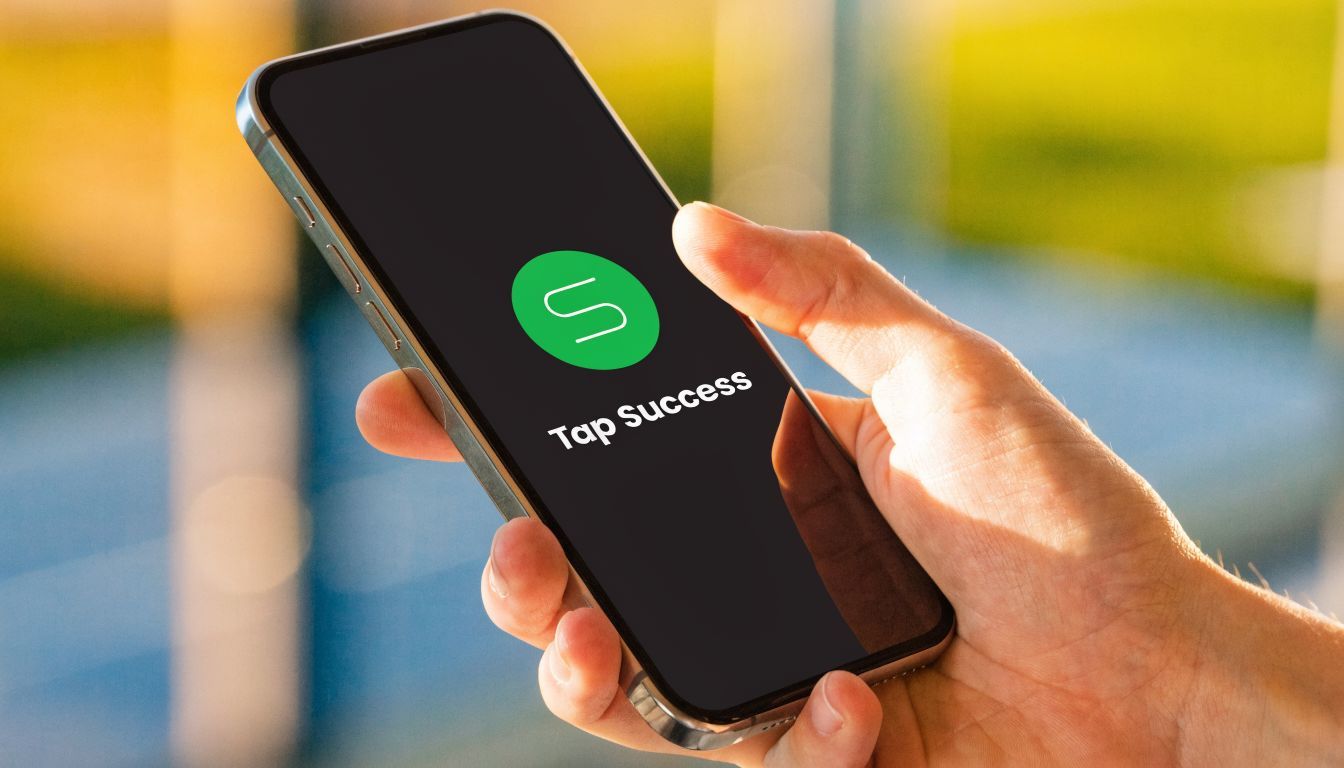 A person holding a smartphone displaying the Tap Success app logo on a blurred outdoor background.