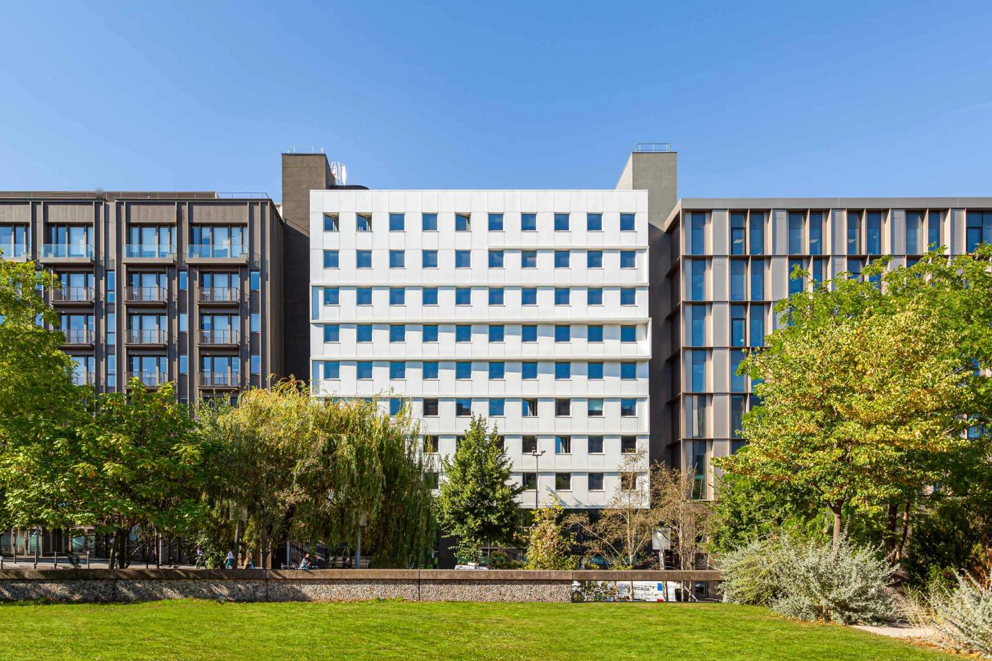Photo of the white-facade Pesaro office building between two dark buildings, lawn in the foreground, Nanterre