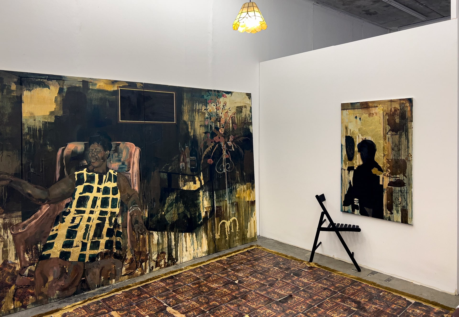 Photograph of a corner of a gallery. On the walls are a large painting of a black woman in an armchair, and a smaller painting featuring a silhouette. Half of a chair is emerging from the wall beneath the painting.