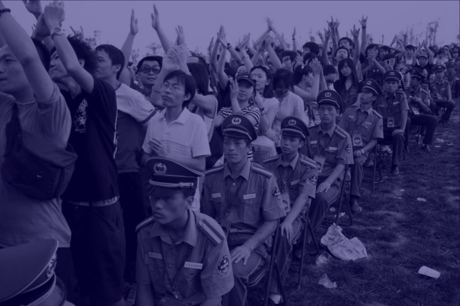 Photograph of a large group of people, facing just to the side of the camera, many with their arms up, and bordered by a row of uniformed police officers sat on foldable chairs.
