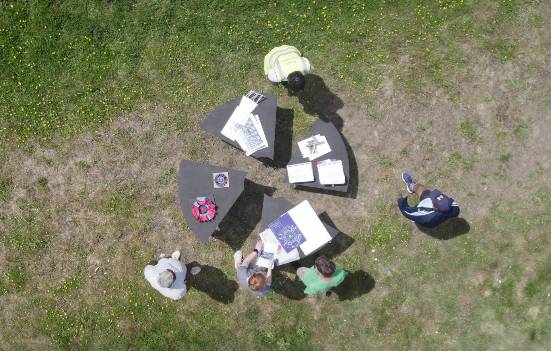 Aerial photograph of four people and four wedge shaped tables of various heights, which have papers on them, in a grassy space.