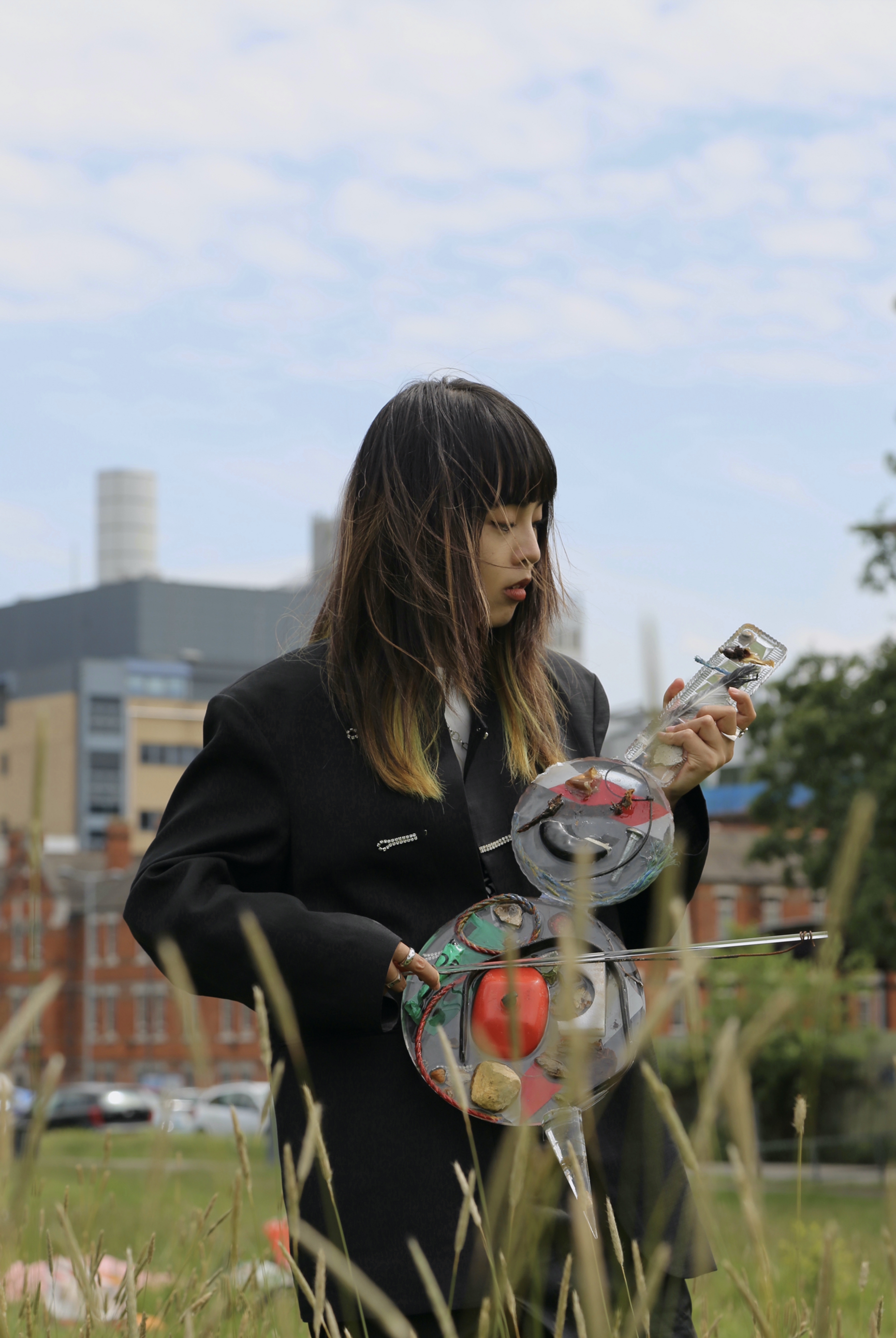 Photograph of a woman from the knees up, standing in a park, with buildings in the distance behind her, playing a musical instrument resembling a large violin made from various materials.