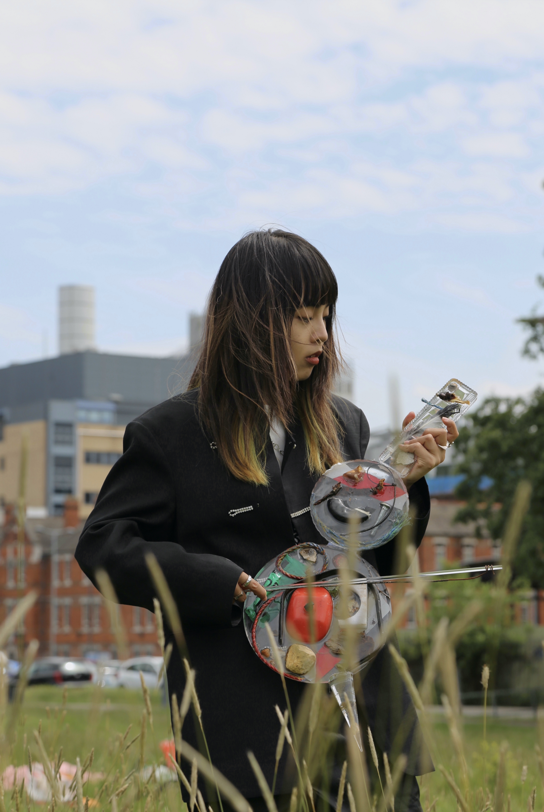 Photograph of a woman from the knees up, standing in a park, with buildings in the distance behind her, playing a musical instrument resembling a large violin made from various materials.