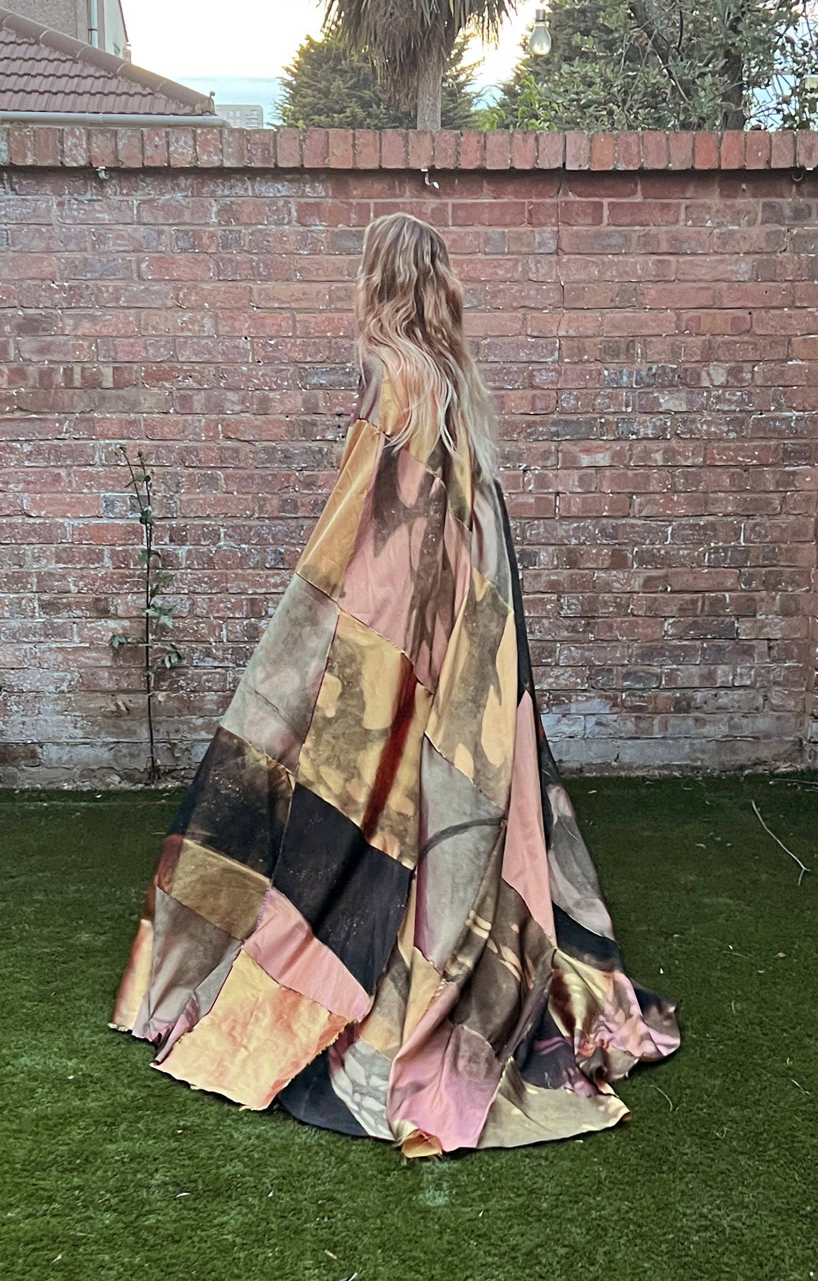 Photograph of a person with their back to the camera, standing on grass in front of a brick garden wall, wearing a long flowing coat or cape made from large squares of different patterned materials.