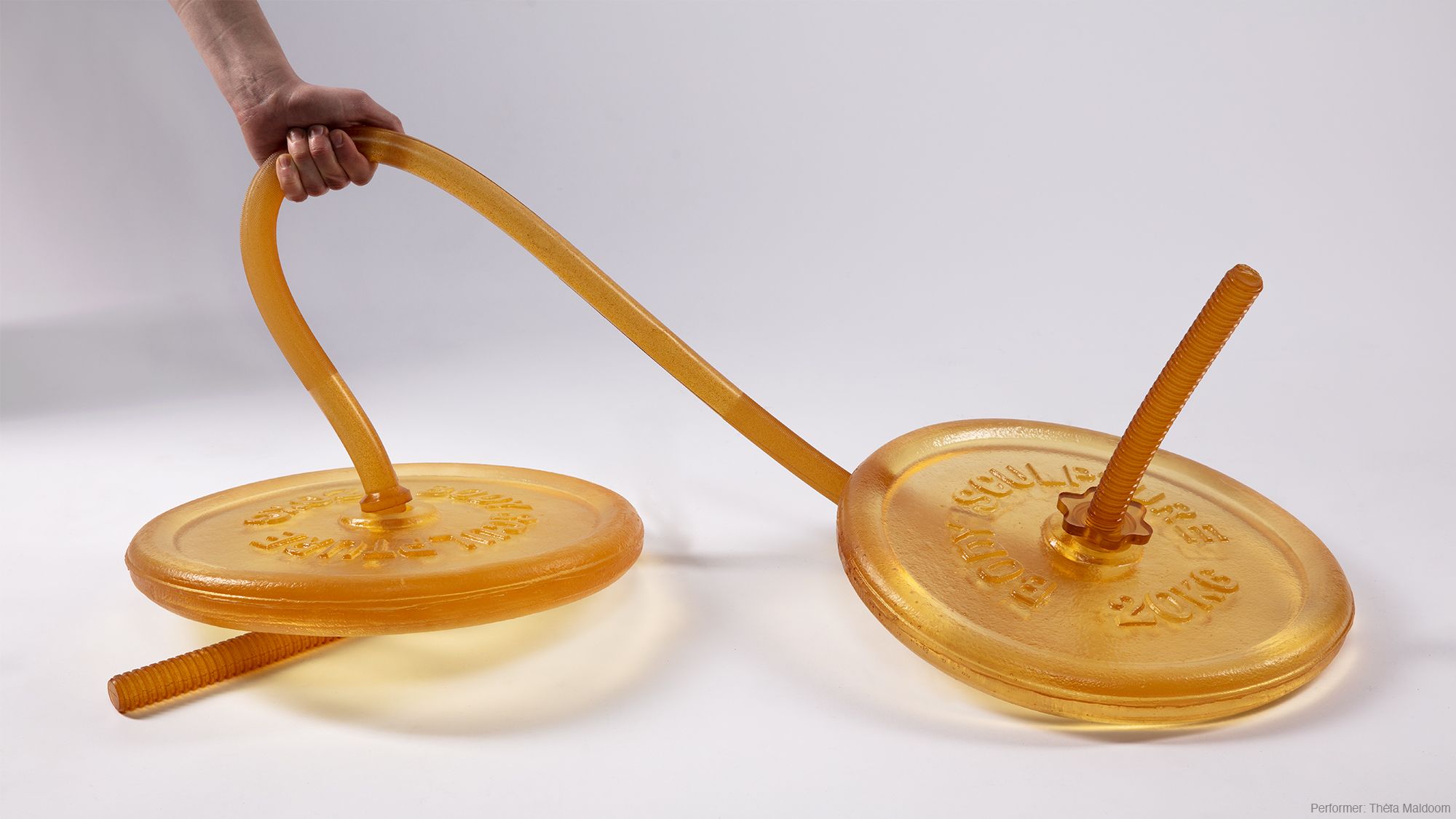 A photograph of a hand reaching down and holding the melted-looking bar of a weight-lifting barbell, which appears to be made from a soft, translucent amber-coloured material. 