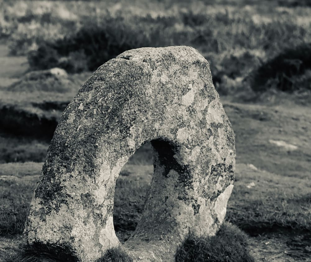 Black and white image, in an outdoor setting, of a ring of stone standing on its side, partly sunk into the ground beneath it, with some tufts grass growing up its base.