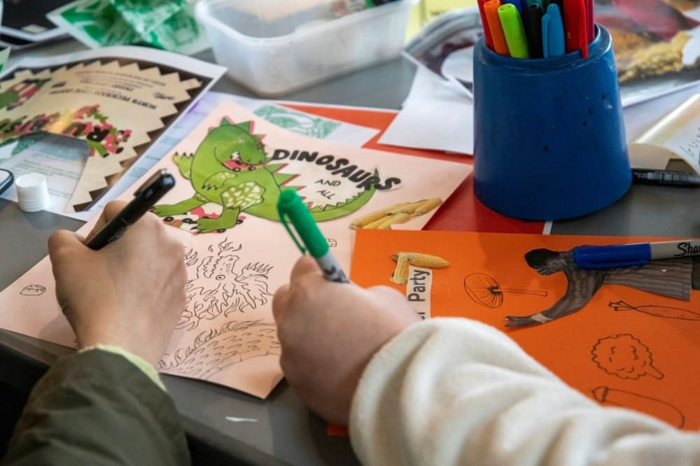 Photograph of two hands holding pens and drawing pictures, one featuring a green dinosaur, on a table-top full of pens and paper.