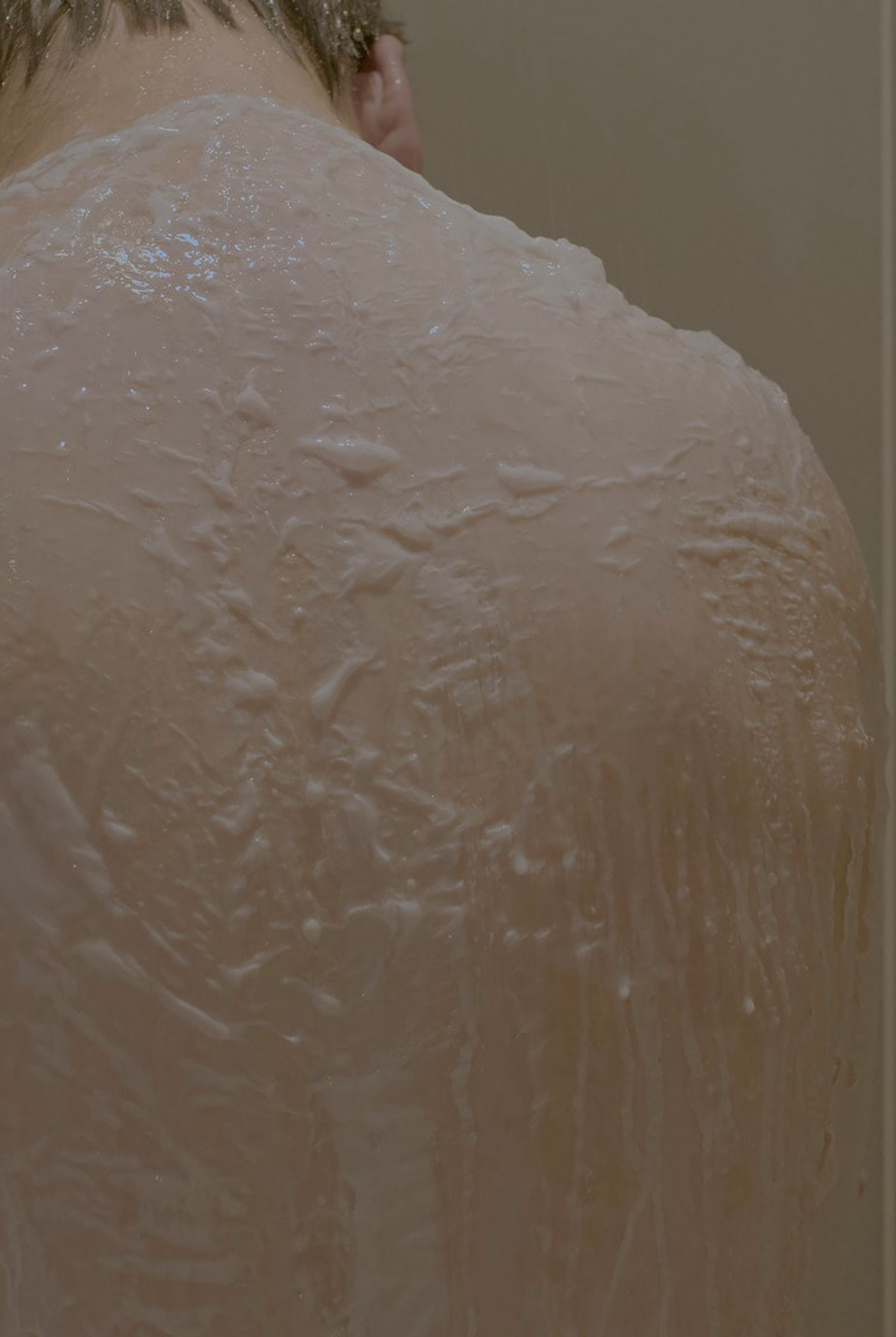 Image of a person with their back to the camera, their head bowed, and their back and upper arms covered in a veil of white soapy foam. By Joe Zijian Zhou