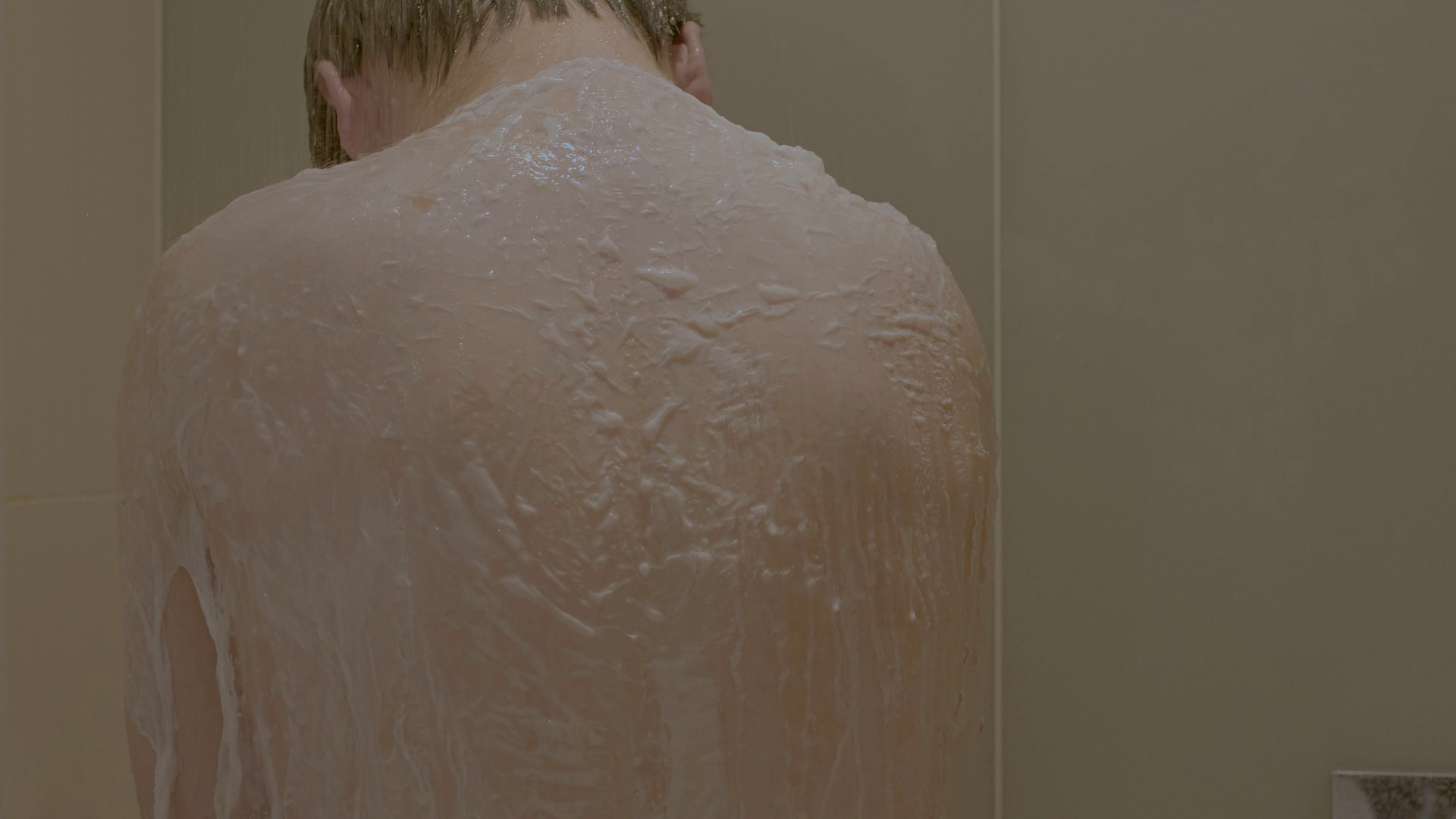 Photograph of the back of a person in a shower, their head bowed, and their back, shoulders and upper arms covered in what appears to be a coating of white soapy foam. 