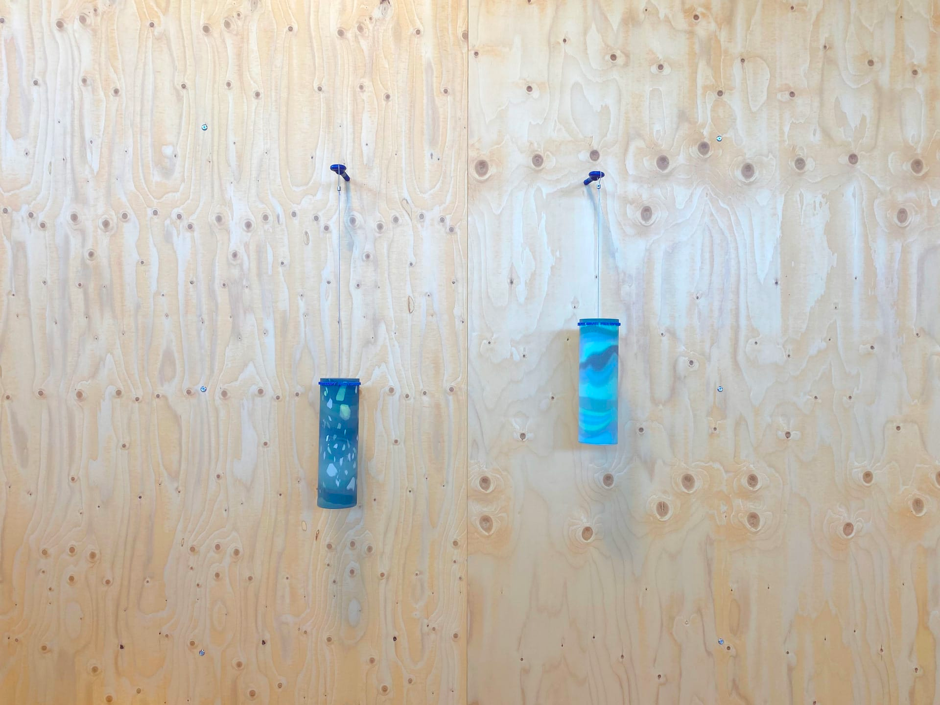 Image of two blue cylindrical objects hanging from string on a wooden wall.