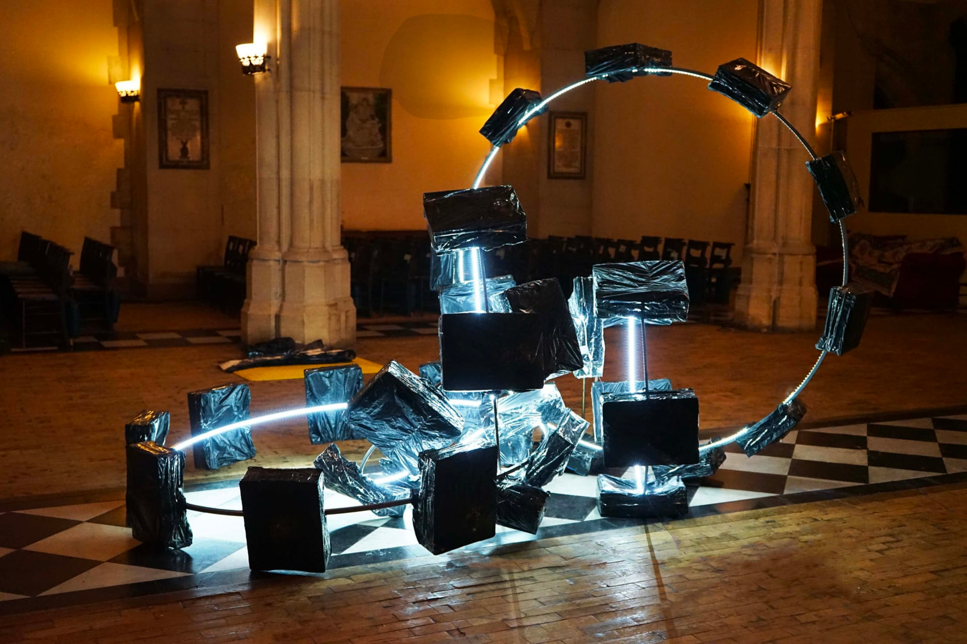 Photograph of a sculpture consisting of several illuminated rings, with black rectangular objects placed around their circumference, in a softly lit room with stone arches and pillars.