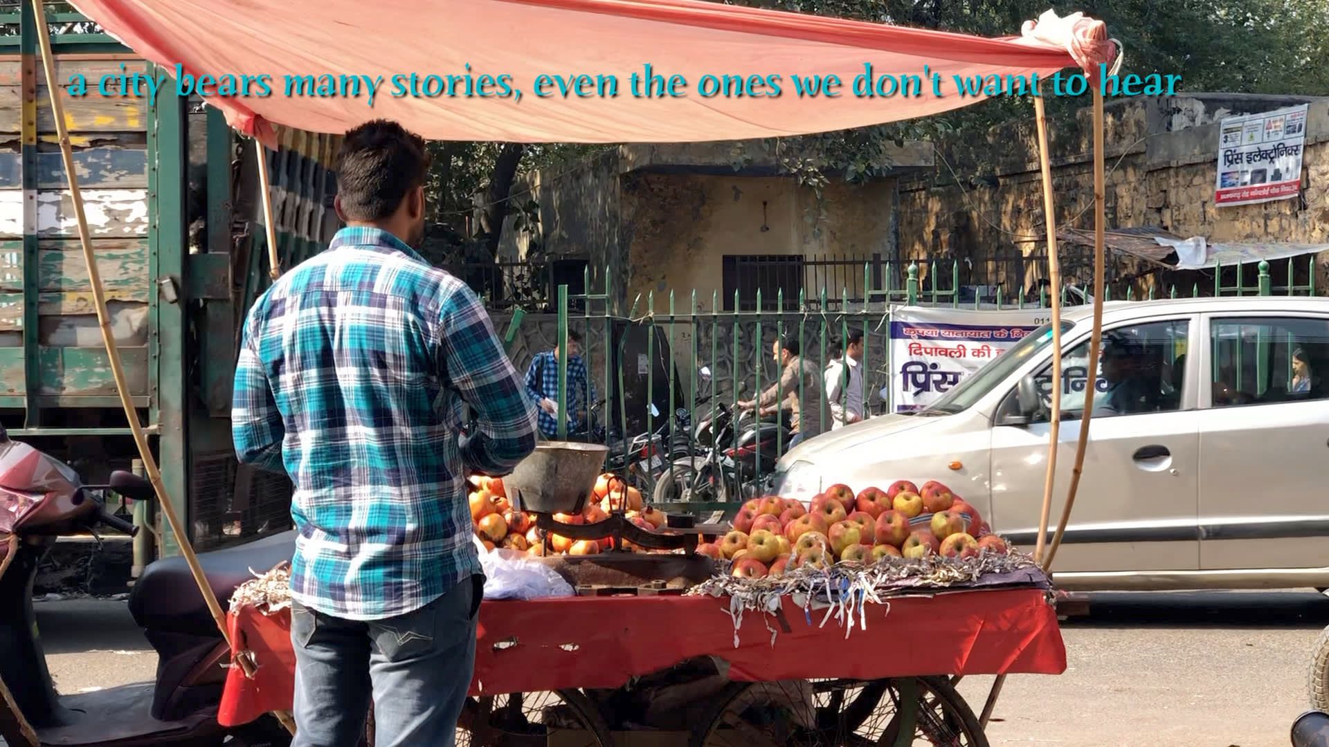 A figure stands with it's back turned in front of an apple cart on the street of an Indian city with the text 'a city bears many stories, even the ones we don't want to hear' across the top.