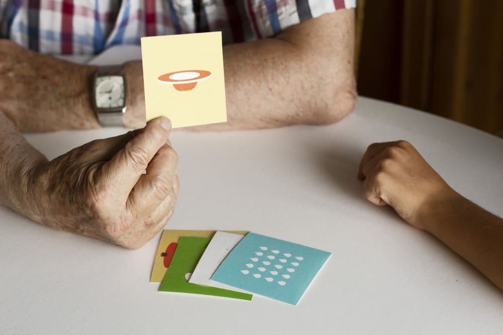 Photograph showing an adult’s arms and hands leaning on a table-top and holding up a card with an image on, with other cards on the table, and a child’s arm resting on the opposite side of the table.