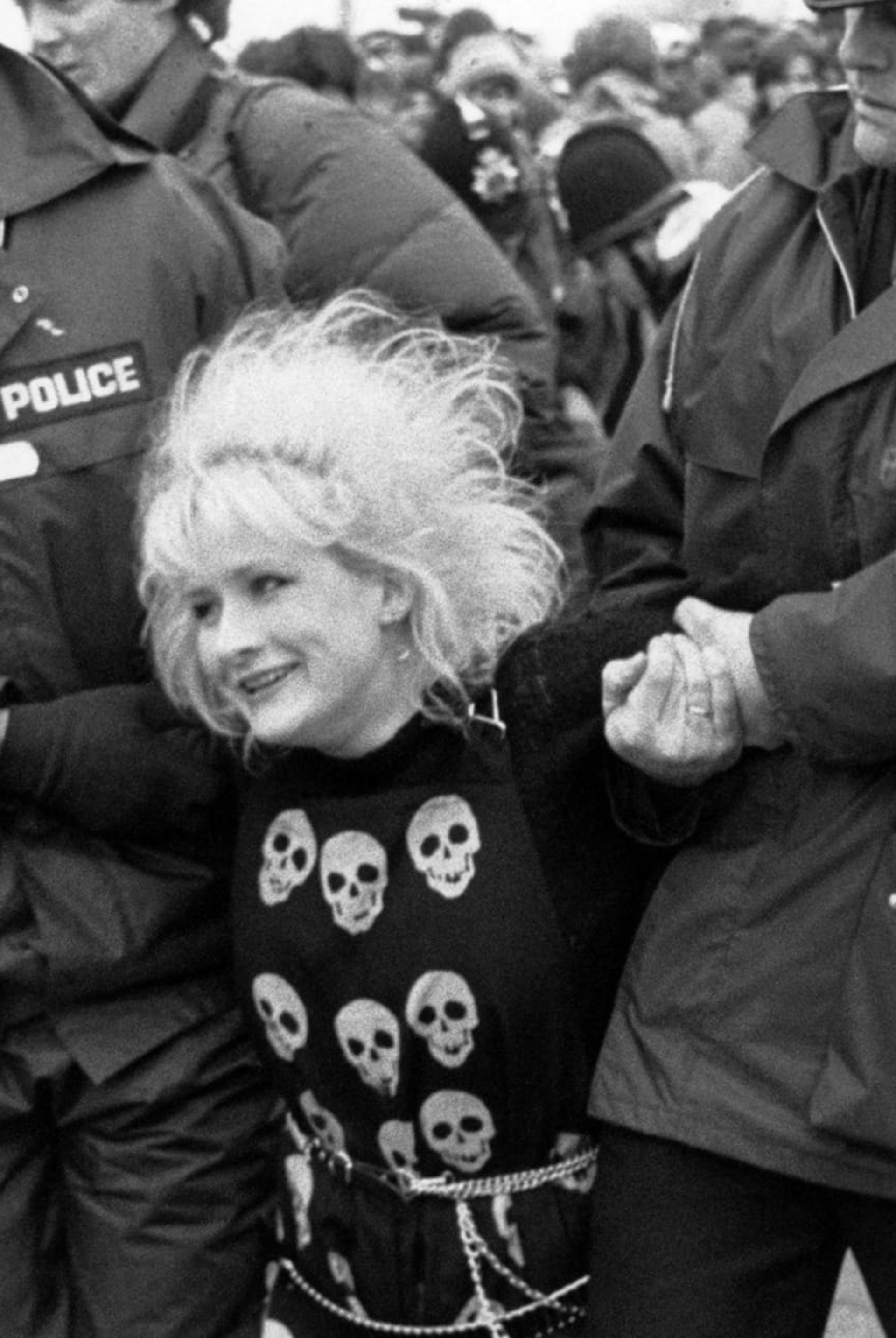 Black and white photograph, which looks as if it was taken in the 1970s or 1980s, of a smiling woman in a top covered in images of skulls, being dragged along by two uniformed British policemen. By Liz Murray