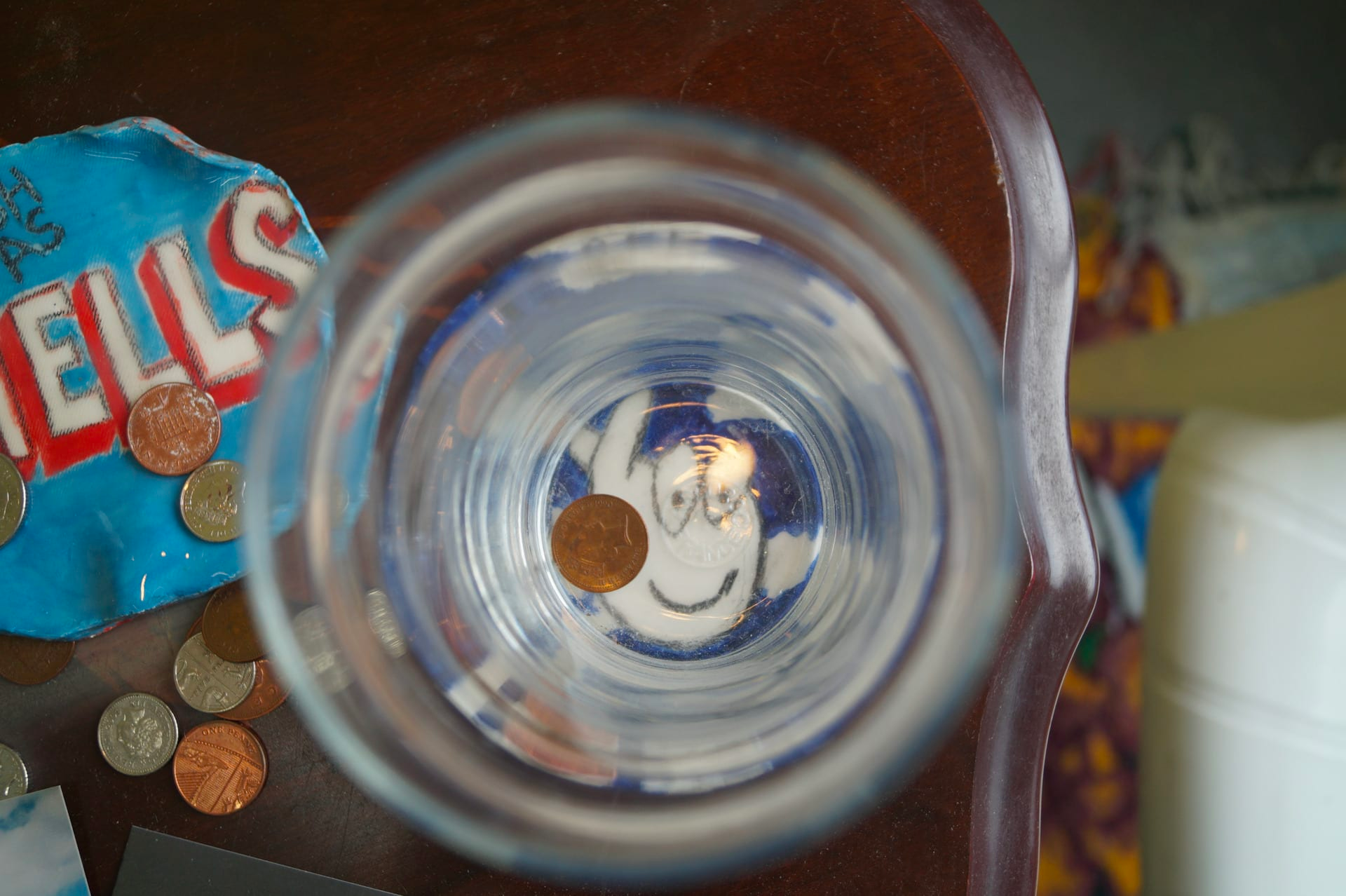 Photograph taken from above, looking into a pint glass, empty except for a small bronze coin, with other coins and objects next to or seen through the glass, which is on the edge of a wooden table.