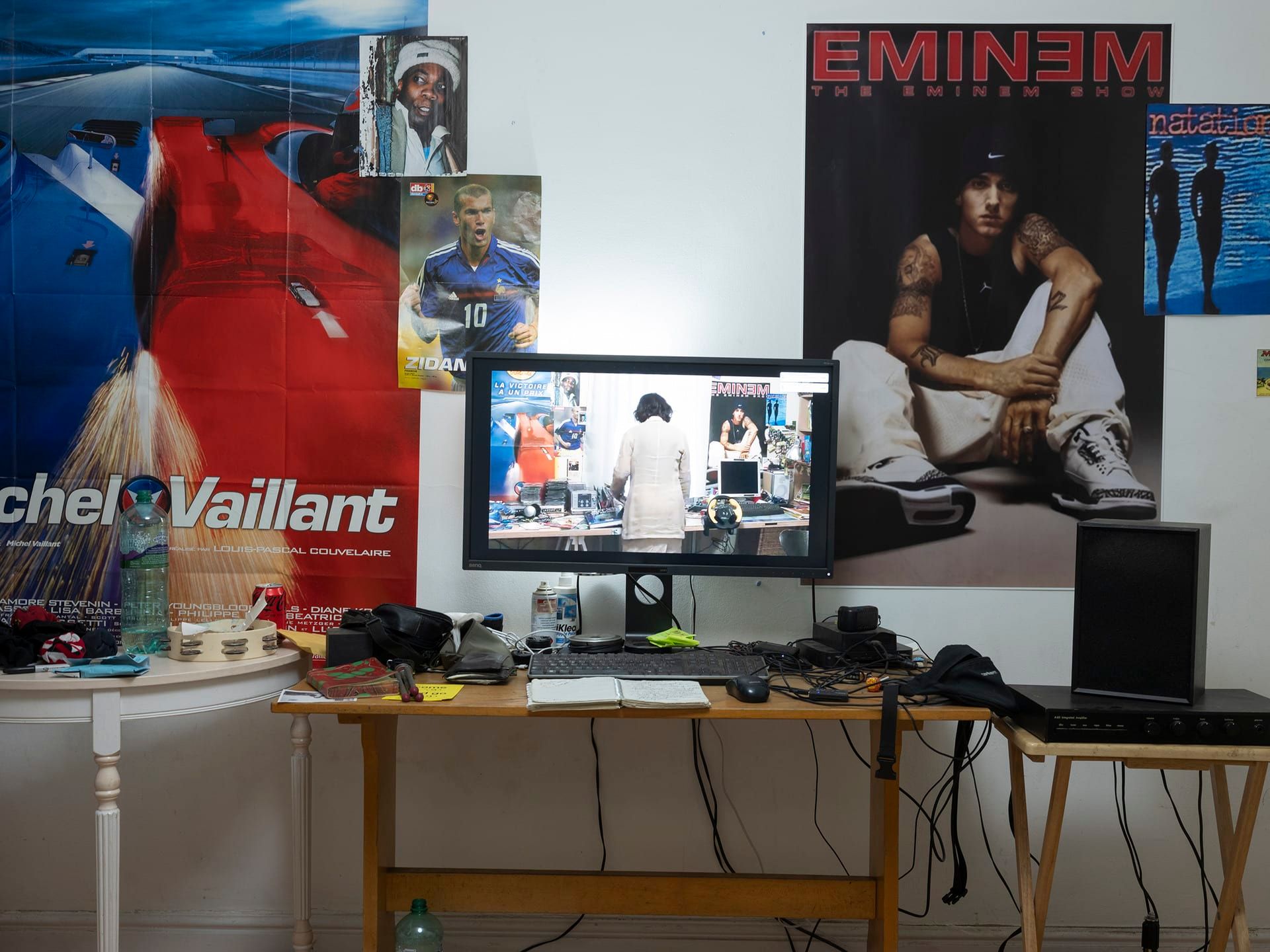 A photo of a series of pop cultural posters sit on a white wall behind a cluttered desk with a computer at the centre displaying the same space but with a figure at the centre turned away from the camera.