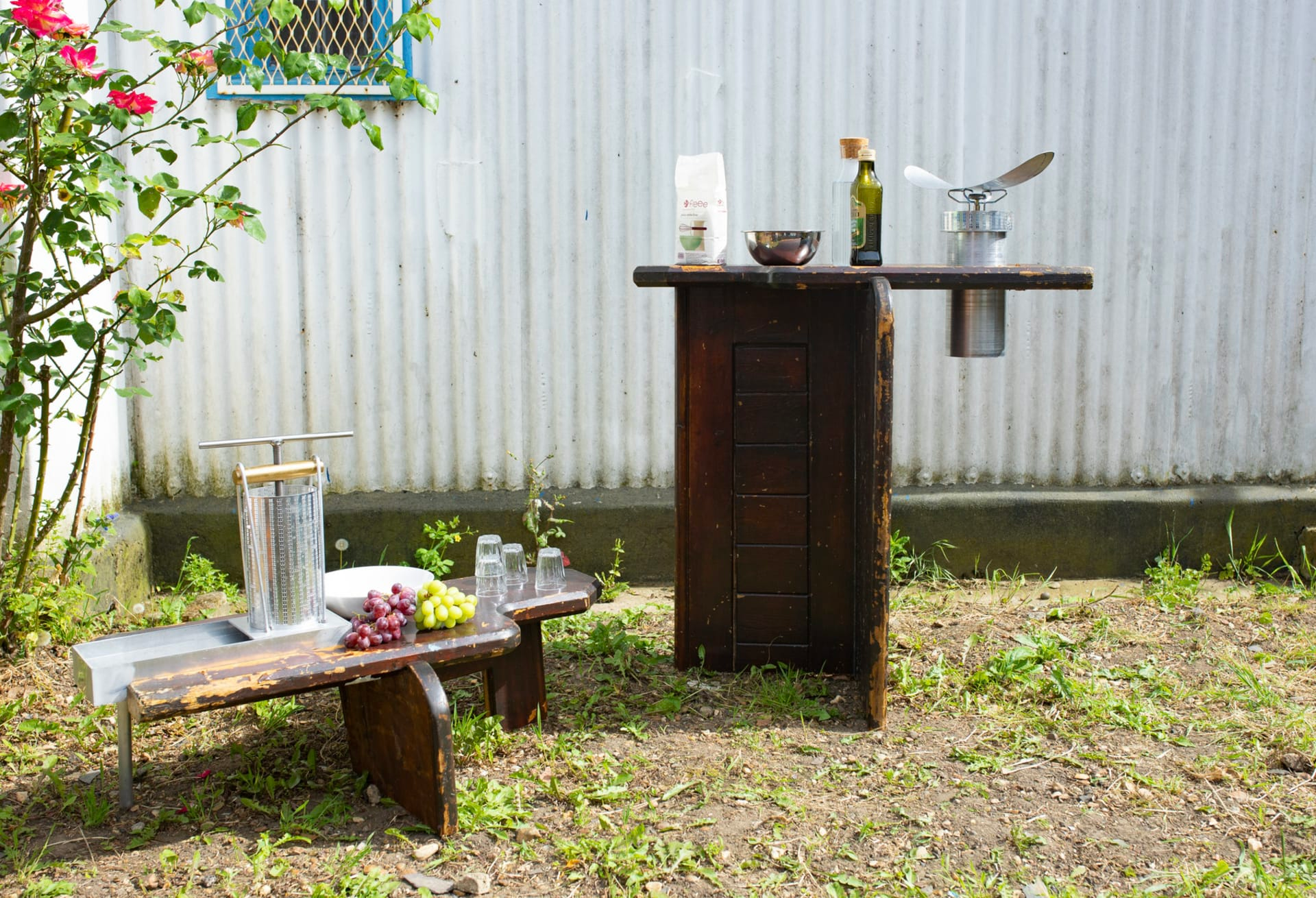 Photograph of two pieces of furniture outside, constructed of shabby dark wood and metal, one a low flat surface holding items, including grapes, the other tall holding a bag of flour and mixing bowl.